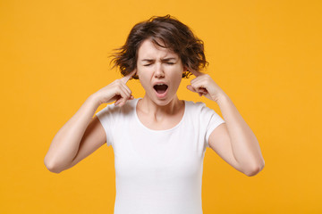 Crazy young brunette woman girl in white t-shirt posing isolated on yellow orange background. People lifestyle concept. Mock up copy space. Covering ears with fingers, keeping eyes closed, screaming.