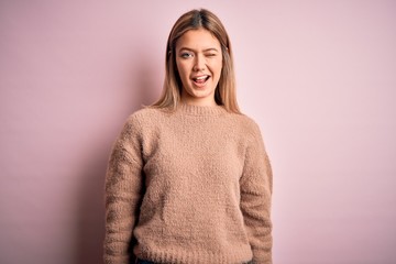 Young beautiful blonde woman wearing winter wool sweater over pink isolated background winking...