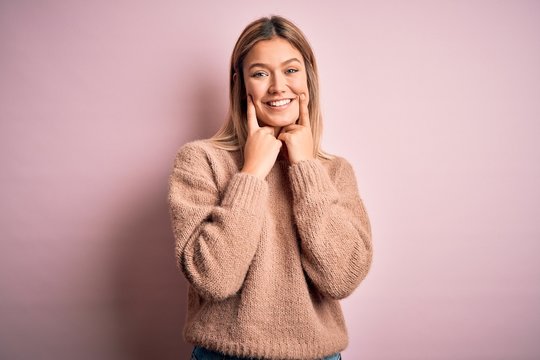 Young beautiful blonde woman wearing winter wool sweater over pink isolated background Smiling with open mouth, fingers pointing and forcing cheerful smile