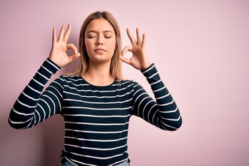 Young beautiful blonde woman wearing casual striped sweater over pink isolated background relax and smiling with eyes closed doing meditation gesture with fingers. Yoga concept.