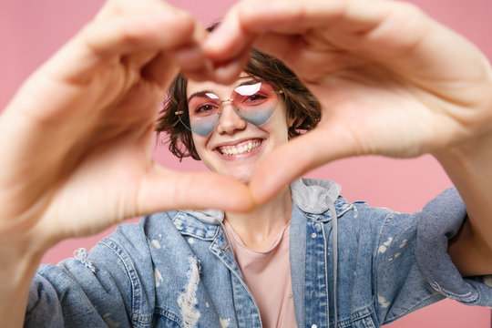 Smiling young brunette woman girl in casual denim jacket, eyeglasses isolated on pastel pink background. People lifestyle concept. Mock up copy space. Showing shape heart with hands, heart-shape sign.