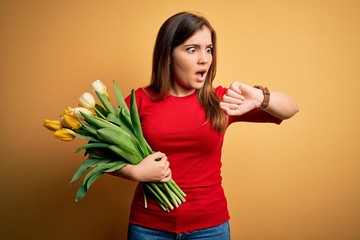 Young blonde woman holding romantic bouquet of tulips flowers over yellow background Looking at the watch time worried, afraid of getting late