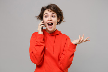 Excited young brunette woman girl in casual red hoodie posing isolated on grey wall background studio portrait. People lifestyle concept. Mock up copy space. Talking on mobile phone, spreading hands.