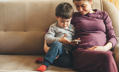 Pregnant caucasian woman with freckles sitting on the sofa with her little boy using a phone
