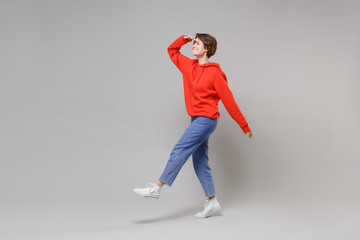 Side view of funny young woman girl in casual red hoodie blue jeans posing isolated on grey background. People lifestyle concept. Mock up copy space. Hold hand at forehead looking far away distance.