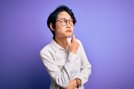 Young beautiful asian girl wearing casual shirt and glasses standing over purple background Thinking worried about a question, concerned and nervous with hand on chin