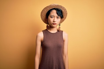Young beautiful asian girl wearing casual t-shirt and hat over isolated yellow background skeptic...