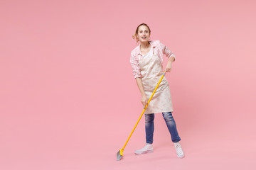 Cheerful funny young woman housewife in casual clothes, apron doing housework isolated on pastel pink wall background studio portrait. Housekeeping concept. Mock up copy space. Sweeping with broom.