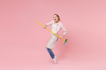 Funny young woman housewife in casual clothes, apron doing housework isolated on pastel pink...