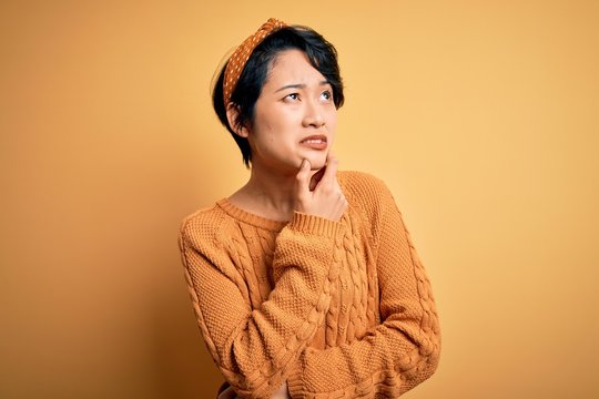 Young beautiful asian girl wearing casual sweater and diadem standing over yellow background Thinking worried about a question, concerned and nervous with hand on chin