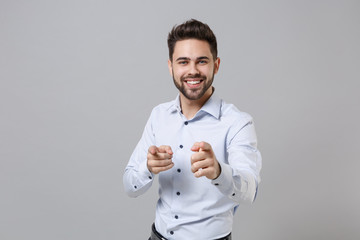 Smiling successful young unshaven business man in light shirt posing isolated on grey wall background. Achievement career wealth business concept. Mock up copy space. Pointing index fingers on camera.