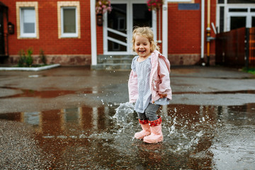 Cute little blonde girl in pink jacket, gray pants and rubber boots is jumping over a puddle on a rainy day