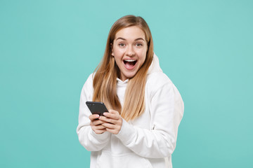 Fototapeta premium Excited young woman girl in casual white hoodie posing isolated on blue turquoise wall background studio portrait. People lifestyle concept. Mock up copy space. Using mobile phone, typing sms message.