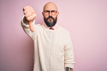 Handsome bald man with beard and tattoo wearing glasses and sweater over pink background looking unhappy and angry showing rejection and negative with thumbs down gesture. Bad expression.