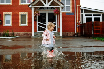 Cute little blonde girl in pink jacket, gray pants and rubber boots is jumping over a puddle on a rainy day