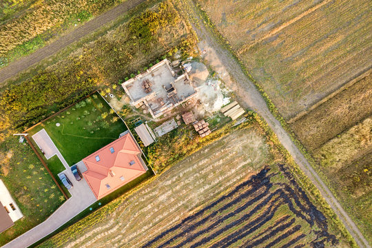 Top down aerial view of two private houses, one under construction with concrete foundament and brick walls and another finished with red tiled roof.