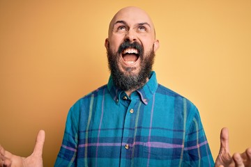 Fototapeta premium Handsome bald man with beard wearing casual shirt standing over isolated yellow background crazy and mad shouting and yelling with aggressive expression and arms raised. Frustration concept.