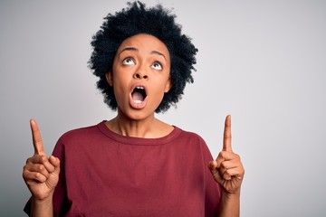 Young beautiful African American afro woman with curly hair wearing casual t-shirt standing amazed and surprised looking up and pointing with fingers and raised arms.