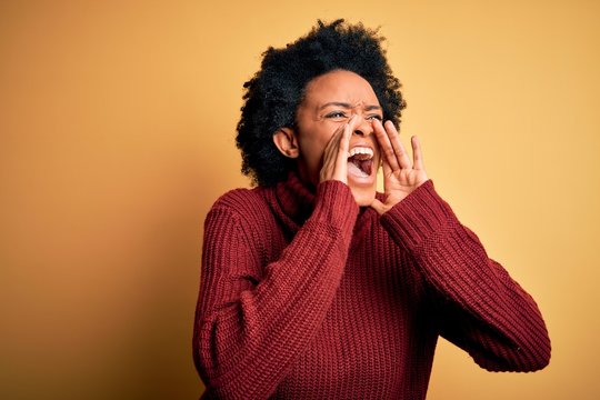 Young beautiful African American afro woman with curly hair wearing casual turtleneck sweater Shouting angry out loud with hands over mouth