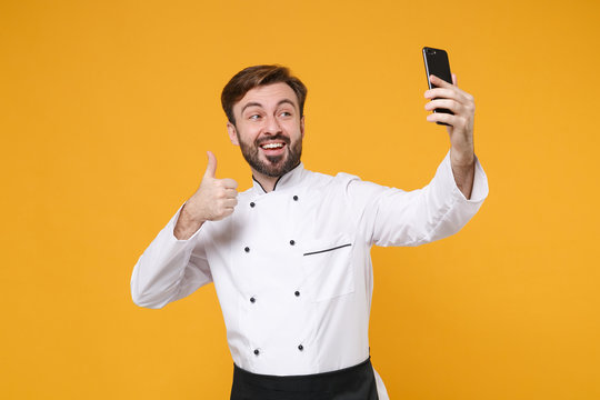 Smiling young bearded male chef cook or baker man in white uniform shirt isolated on yellow background. Cooking food concept. Mock up copy space. Doing selfie shot on mobile phone, showing thumb up.