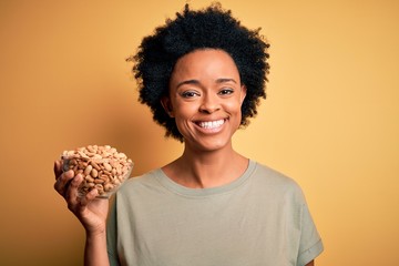 Young African American afro woman with curly hair holding bowl with salty peanuts with a happy face standing and smiling with a confident smile showing teeth
