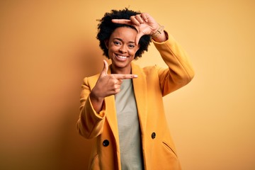 Young beautiful African American afro businesswoman with curly hair wearing yellow jacket smiling making frame with hands and fingers with happy face. Creativity and photography concept.