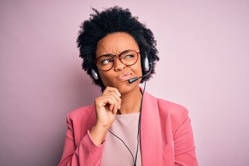 Young African American call center operator woman with curly hair using headset with hand on chin...