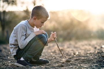 Child boy playing with wooden stick digging in black dirt ground outdoors.