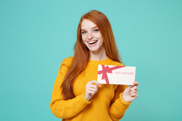 Cheerful young redhead woman girl in yellow knitted sweater posing isolated on blue turquoise background studio portrait. People emotions lifestyle concept. Mock up copy space. Hold gift certificate.