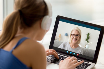 View over shoulder of adult daughter talks by video call with 50s mum. Pc screen view smiling...