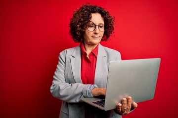 Middle age curly hair business woman working using laptop over isolated red background with a confident expression on smart face thinking serious