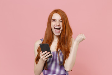 Happy young redhead woman girl in plaid dress posing isolated on pastel pink background studio portrait. People emotions lifestyle concept. Mock up copy space. Using mobile phone doing winner gesture.