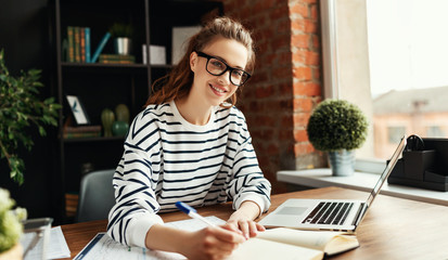 Happy millennial woman taking notes in notepad while working at laptop in comfortable loft office.