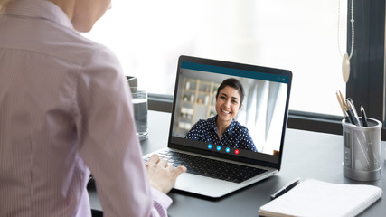 Indian girl communicate with friend on-line by video call, pc screen view over female shoulder. Mental health expert online therapy, colleagues work on common project use videoconferencing app concept - obrazy, fototapety, plakaty