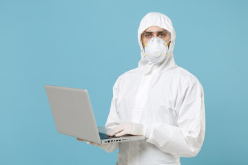 Man in white protective suit respirator mask hold using laptop pc computer isolated on blue background studio. Epidemic pandemic new rapidly spreading coronavirus 2019-ncov medicine flu virus concept.
