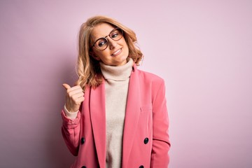 Middle age beautiful blonde business woman wearing elegant pink jacket and glasses smiling with happy face looking and pointing to the side with thumb up.