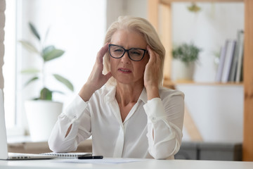 Unhappy 60 years old businesswoman massaging temples close up, suffering from strong head ache. Exhausted stressful employee feeling pain or blood pressure, unwell, health problem concept.
