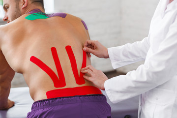 Kinesiology taping. Physical therapist applying kinesiology tape to patient back. Therapist treating injured lower back of young male athlete. Post traumatic rehabilitation, sport physical therapy.