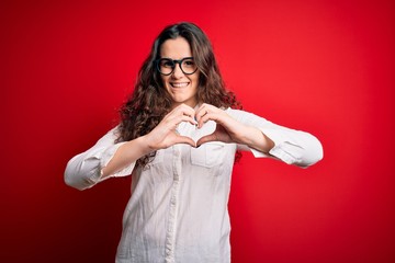 Young beautiful woman with curly hair wearing shirt and glasses over red background smiling in love showing heart symbol and shape with hands. Romantic concept.