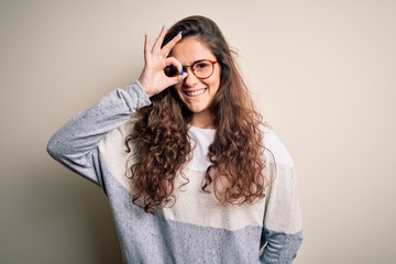 Young beautiful woman with curly hair wearing sweater and glasses over white background doing ok gesture with hand smiling, eye looking through fingers with happy face.