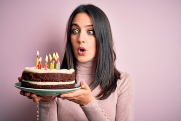 Young woman with blue eyes holding birthday cake with candles over pink background scared in shock...
