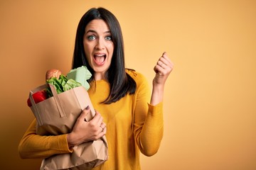 Young brunette woman with blue eyes holding healthy groceries in paper bag from supermarket screaming proud and celebrating victory and success very excited, cheering emotion