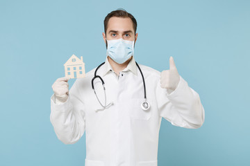Doctor man in medical gown face mask gloves isolated on blue background. Epidemic pandemic coronavirus 2019-ncov sars covid-19 flu virus concept. Hold house recommending staying home showing thumb up.