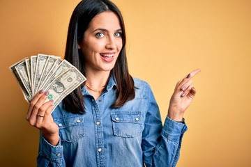 Young brunette woman with blue eyes holding 1 dollar banknotes over yellow isolated background very happy pointing with hand and finger to the side