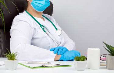 doctor in a white coat and blue latex gloves sits at a white work table in his office