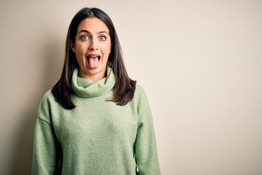 Young brunette woman with blue eyes wearing turtleneck sweater over white background sticking tongue out happy with funny expression. Emotion concept.