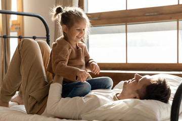 Fototapeta premium Full length happy young father lying on comfortable bed, holding on belly small preschool kid daughter. Cheerful single dad having fun playing together with cute little child girl in bedroom.
