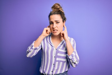 Young beautiful blonde woman wearing casual striped shirt standing over purple background covering ears with fingers with annoyed expression for the noise of loud music. Deaf concept.