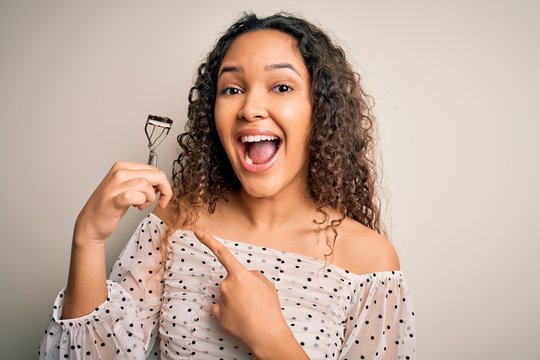 Young beautiful woman with curly hair holding eyelases curler over white background very happy pointing with hand and finger