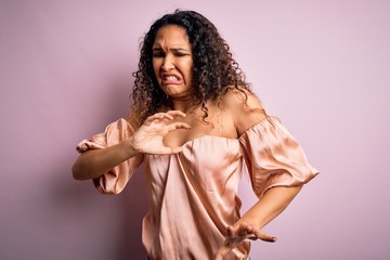 Young beautiful woman with curly hair wearing casual t-shirt standing over pink background disgusted expression, displeased and fearful doing disgust face because aversion reaction.